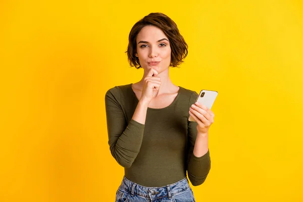 Foto di cute ragazza tenere braccio smartphone mentalità guardare fotocamera indossare camicia verde isolato luminoso colore giallo sfondo — Foto Stock