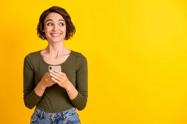 Foto de menina linda segurar celular olhar lado espaço vazio dente sorriso desgaste camisa verde isolado vívido cor amarela fundo — Fotografia de Stock