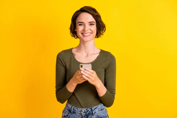 Foto de encantadora joven dama sostener teléfono mirar cámara brillante sonrisa usar camisa verde aislado vibrante color amarillo fondo —  Fotos de Stock