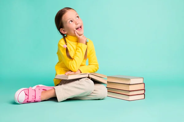 Foto de tamaño completo de pelo castaño niña elegante sentarse piso dedo mejilla mirada espacio vacío usar pantalones blancos cuello alto amarillo aislado sobre fondo de color verde azulado — Foto de Stock