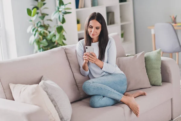 Foto de cuerpo completo de la calma encantadora morena joven mujer sentarse sofá usar jeans usar teléfono dentro de casa en interiores —  Fotos de Stock