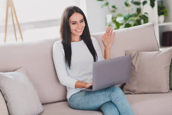 Foto av charmig positiv kvinna vinka hand säga hej video på laptop sitta soffa hem inomhus i vardagsrummet — Stockfoto