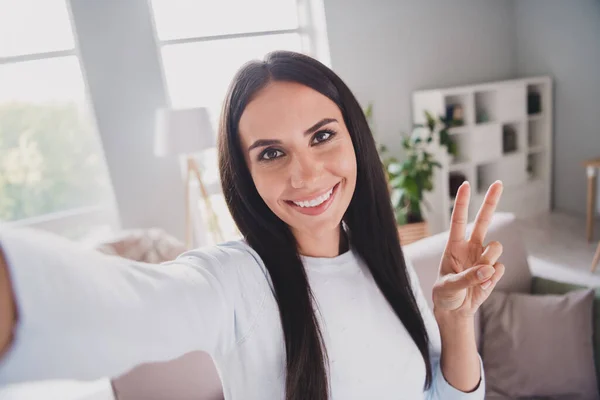 Фото счастливой веселой привлекательной молодой женщины сделать фотоаппарат селфи в гостиной показать V-знак в помещении в доме — стоковое фото