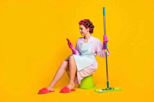 Teljes méretű fotó lány mosás padló ülés vödör használata okostelefon kopás köntös kesztyű elszigetelt sárga szín háttér — Stock Fotó