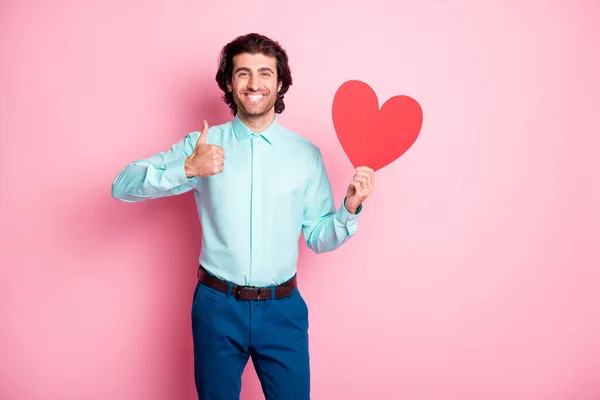 Photo portrait of man holding red heart card showing thumb up isolated on pastel pink colored background — Stock Photo, Image