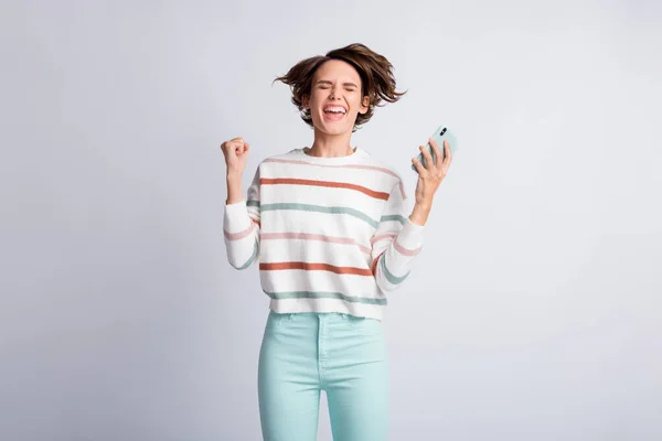 Foto de mujer joven afortunada vestida con un jersey a rayas sosteniendo un gadget moderno levantando los ojos cerrados con puño aislado color gris fondo — Foto de Stock