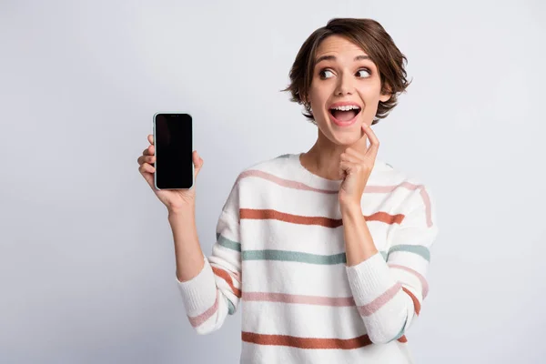 Foto de mujer joven linda sorprendida vestida con jersey a rayas que sostiene el dispositivo moderno barbilla de dedo aislado fondo de color gris —  Fotos de Stock