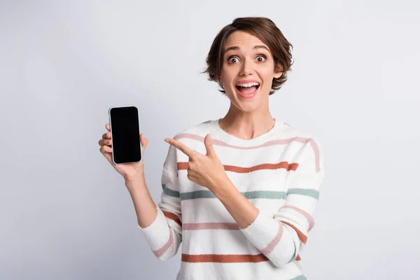 Foto de mujer joven excitada usar suéter rayado apuntando con la celebración de la boca abierta gadget moderno aislado fondo de color gris —  Fotos de Stock
