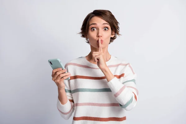 Foto de señora joven divertida usar suéter a rayas que sostiene gadget moderno pedir no contar secretos aislados fondo de color gris — Foto de Stock