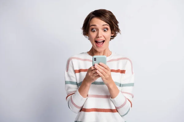 Foto de mujer joven emocionada impresionada vestida con un pulóver a rayas que sostiene un dispositivo moderno aislado fondo de color gris —  Fotos de Stock
