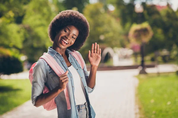 Foto porträtt av svart flådd söt flicka med lockigt hår leende hälsning viftande hand hålla ryggsäck bär denim outfit — Stockfoto