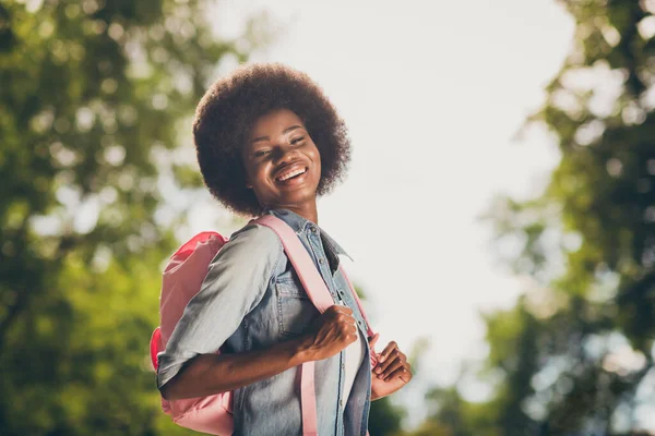 Boczny profil zdjęcie czarnej skóry ładna dziewczyna z kręconymi włosami śmiejąc się szczęśliwie utrzymując plecak uśmiechnięty w zielonym parku miejskim — Zdjęcie stockowe