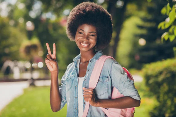 Foto porträtt av svarthyade lockigt flicka ler glatt visar v-tecken med två fingrar bär denim skjorta rosa casual ryggsäck — Stockfoto