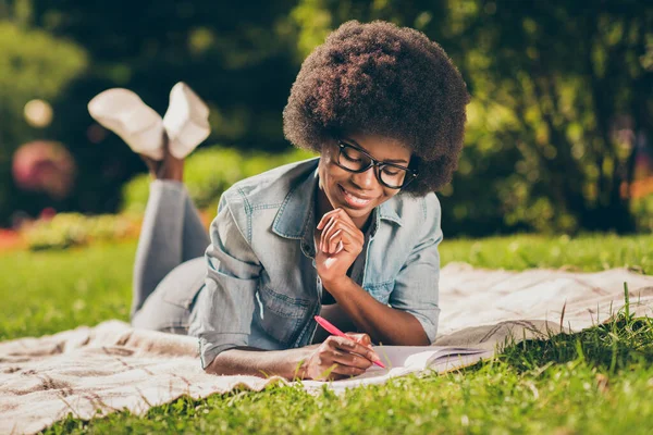 Ritratto foto di sognante sorridente ragazza dalla pelle nera sdraiata nel parco, studiando scrittura saggio in copybook indossa acconciatura riccia — Foto Stock