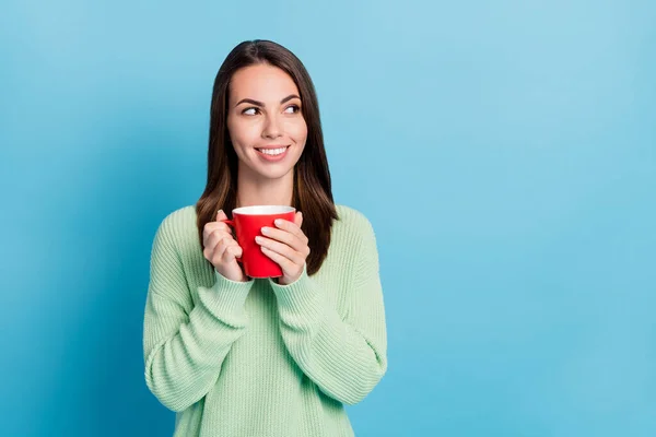 Foto retrato de menina com cabelo morena segurando xícara de bebida quente sorrindo olhando para o lado vestindo roupas verdes isoladas em fundo de cor azul vibrante com espaço em branco — Fotografia de Stock