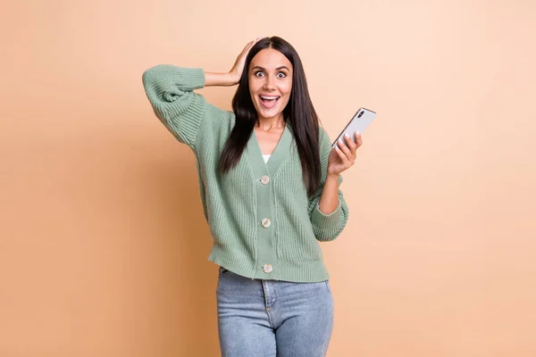 Foto van onder de indruk schattig meisje houden telefoon hand haar dragen groene trui jeans geïsoleerd op perzik kleur achtergrond — Stockfoto