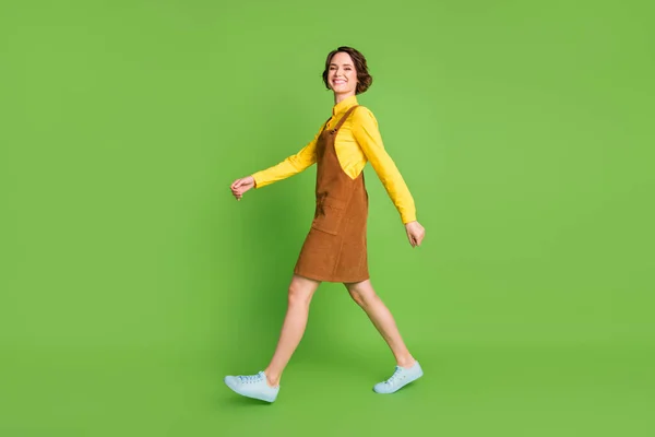Full längd profil foto av gående flicka utseende kamera bära gul skjorta övergripande skor isolerad grön färg bakgrund — Stockfoto