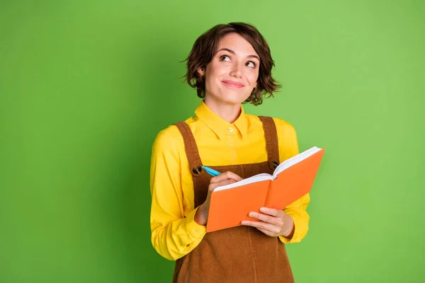 Foto di sognante giovane ragazza sguardo vuoto spazio tenere copybook penna scrivere indossare camicia gialla complessiva isolato colore verde sfondo — Foto Stock