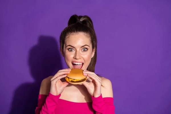 Közelkép portré vonzó vidám lány eszik friss forró burger harapás elszigetelt felett fényes lila színű háttér — Stock Fotó