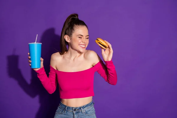 Porträtt av attraktiva smal glad flicka äta hamburgare dricka cola njuta skrattar isolerad över ljusa violett färg bakgrund — Stockfoto