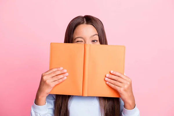 Primer plano retrato de linda linda chica de pelo castaño divertido escondido detrás de libro guiño aislado sobre fondo de color pastel rosa — Foto de Stock