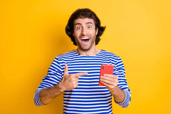 Foto van verbaasde gekke man wijzen vinger smartphone dragen blauw wit vest geïsoleerd over helder gele kleur achtergrond — Stockfoto