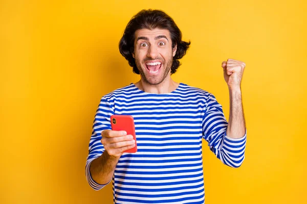 Foto van extatische man houden smartphone verhogen vuisten schreeuwen dragen blauw wit shirt geïsoleerd op glans geel kleur achtergrond — Stockfoto