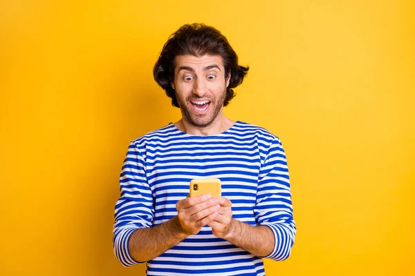 Hayret bir adamın fotoğrafı akıllı telefondaki sosyal medya paylaşımlarını okuyor parlak sarı arka planda izole edilmiş beyaz mavi gömlek giyiyor — Stok fotoğraf