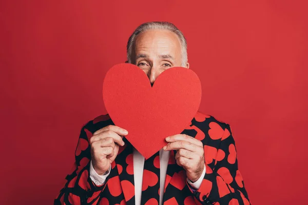 Photo of shy retired old man hold paper heart figure closing face look camera wear suit isolated red background