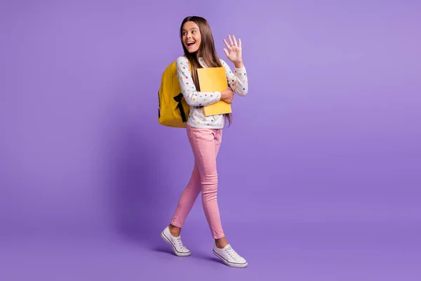 Tamanho total do corpo foto de aluno morena indo para a escola abraçando livro carregando mochila acenando isolado em fundo violeta vívido — Fotografia de Stock