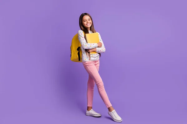 Tam boy beden ölçülerinde, kitap taşıyan, pembe pantolon giyen parlak mor arka planda izole edilmiş kazak giyen bir kızın fotoğrafı. — Stok fotoğraf
