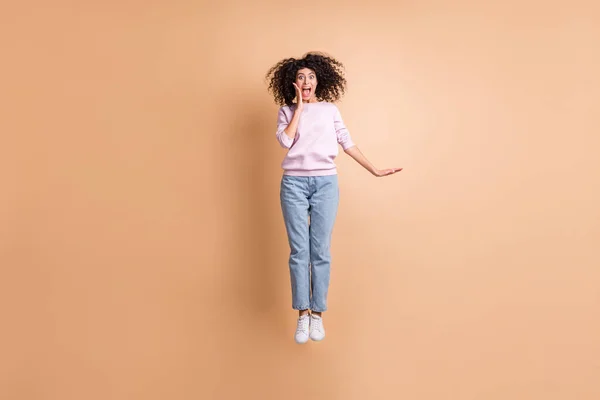 Foto de tamaño completo de salto impresionado chica rizada usar suéter jeans zapatillas aisladas en el fondo de color melocotón —  Fotos de Stock