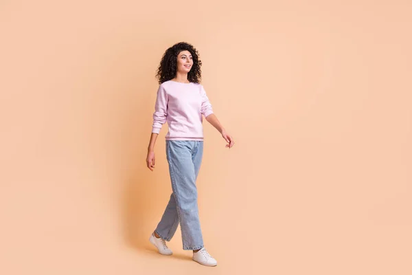 Foto de tamaño completo de optimista ir chica rizada usar suéter jeans zapatillas aisladas en el fondo de color melocotón —  Fotos de Stock