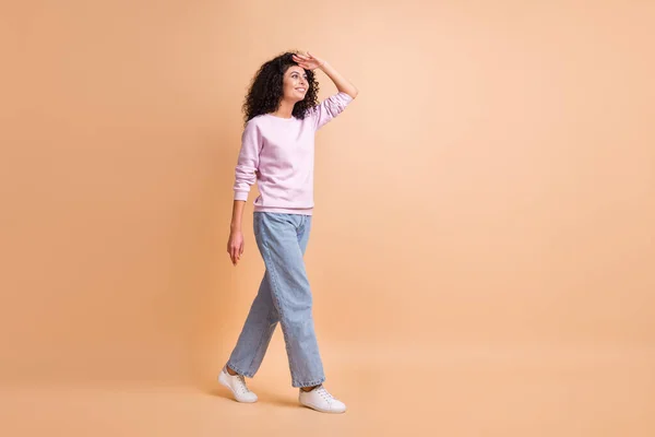 Foto de tamaño completo de optimista ir chica rizada ver a alguien usar suéter jeans zapatillas aisladas en el fondo de color melocotón —  Fotos de Stock