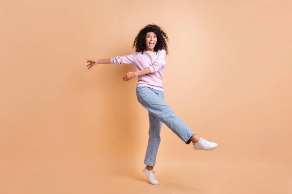 Foto de tamaño completo de optimista chica rizada bailando desgaste suéter jeans zapatillas aisladas en el fondo de color melocotón —  Fotos de Stock