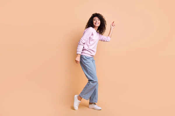 Foto de perfil de tamaño completo de optimista chica rizada bailando usar suéter jeans zapatillas aisladas en el fondo de color melocotón —  Fotos de Stock