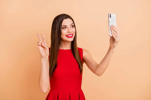 Çekici, mutlu bir kadının fotoğrafı. V işaretli telefon al, selfie çek. Bej renkli arka planda izole edilmiş kırmızı elbise giy. — Stok fotoğraf