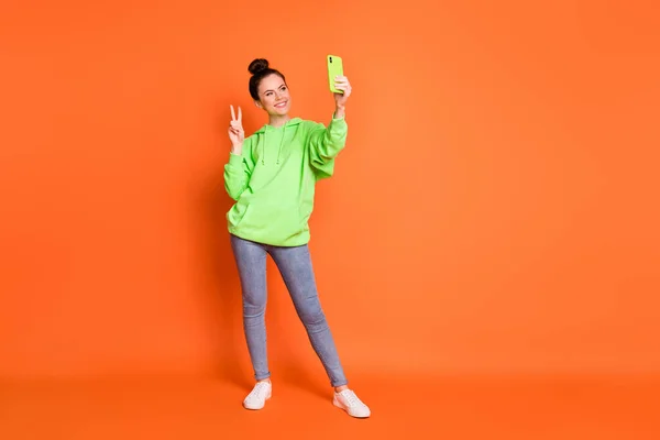Foto ritratto vista corpo pieno di ragazza carina mostrando v-segno che tiene il telefono in una mano prendendo selfie isolato su vivido sfondo di colore arancione con spazio vuoto — Foto Stock
