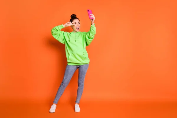 Retrato de foto de comprimento total da menina fazendo v-sinal perto do olho segurando telefone em uma mão rindo tirando selfie isolado no fundo colorido laranja vívido — Fotografia de Stock