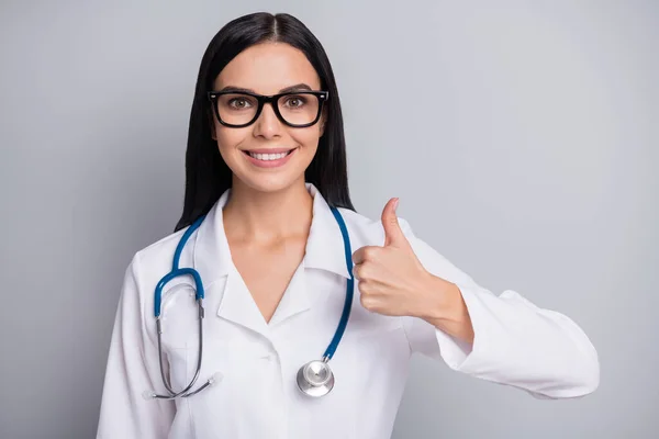Foto de mujer terapeuta positiva alegre vestida con gafas uniformes blancas que muestran el pulgar hacia arriba fondo de color gris aislado — Foto de Stock