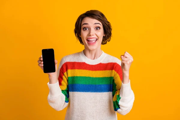 Foto van verbaasd dame houden telefoon tonen touchscreen lege ruimte verhogen vuist slijtage gestreepte pullover geïsoleerde gele kleur achtergrond — Stockfoto