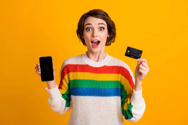 Фото милой леди держать дебетовую карту телефон показывают пустое пространство с открытым ртом носить полосатый пуловер изолированный желтый цвет фона — стоковое фото