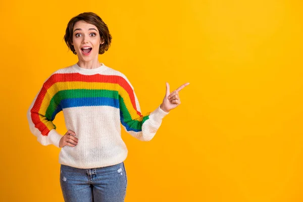 Photo of optimistic surprised brunette hair girl stand point empty space wear rainbow sweater hand waist isolated on yellow color background