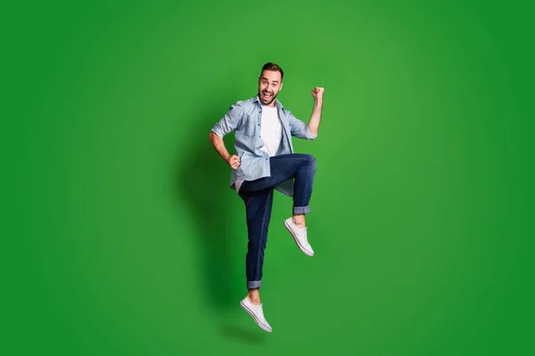 Full storlek profil foto av imponerad rolig kille hoppa bära blå skjorta jeans skor isolerade på grön färg bakgrund — Stockfoto