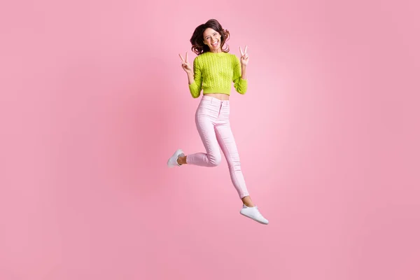 Pleine longueur photo de charmante femme bouclée habillée pullover vert saut montrant v-signe isolé fond de couleur rose — Photo