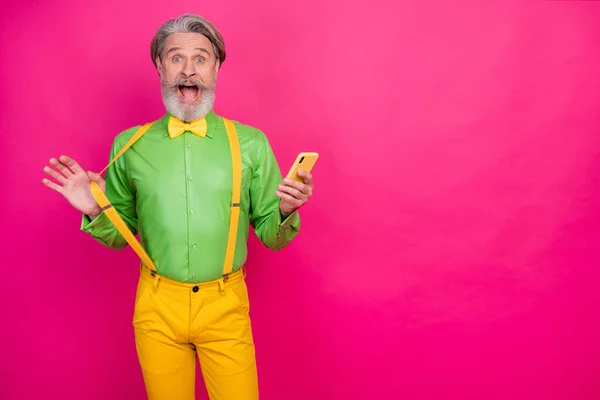 Fénykép funky idős nagypapa nyitott száj ideális bajusz tartsa telefon tetteti millenniumi viselet zöld ing sárga nadrágtartó csokornyakkendő nadrág elszigetelt fényes rózsaszín háttér — Stock Fotó