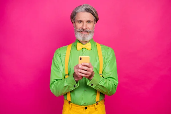 素敵な白い髪のおじいちゃんの写真を保持電話ブログ記事現代的な技術のユーザーは緑のシャツを着用黄色のサスペンダー弓ネクタイパンツ孤立明るいピンク色の背景 — ストック写真