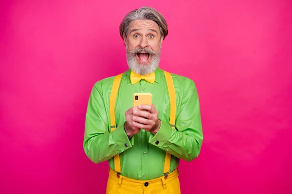Şaşırmış beyaz saçlı büyükbabanın fotoğrafı telefon tut blog yorumları oku yeşil gömlek giy sarı pantolon giy parlak pembe arka plan — Stok fotoğraf