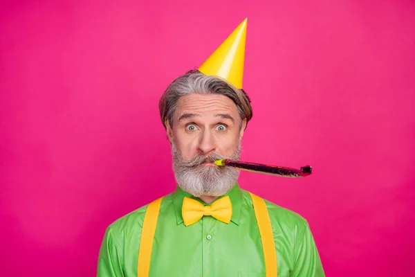 Fénykép a vicces fehér hajú nagypapa fúj zajongó funky születésnapi party hűtés kopás papír sapka zöld ing sárga nadrágtartó elszigetelt élénk rózsaszín háttér — Stock Fotó