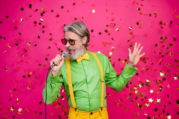 Foto av roliga galna morfar klubbfest kyla sång karaoke använda mikrofon konfetti falla bära sol specs grön skjorta gula hängslen isolerad ljus rosa färg bakgrund — Stockfoto